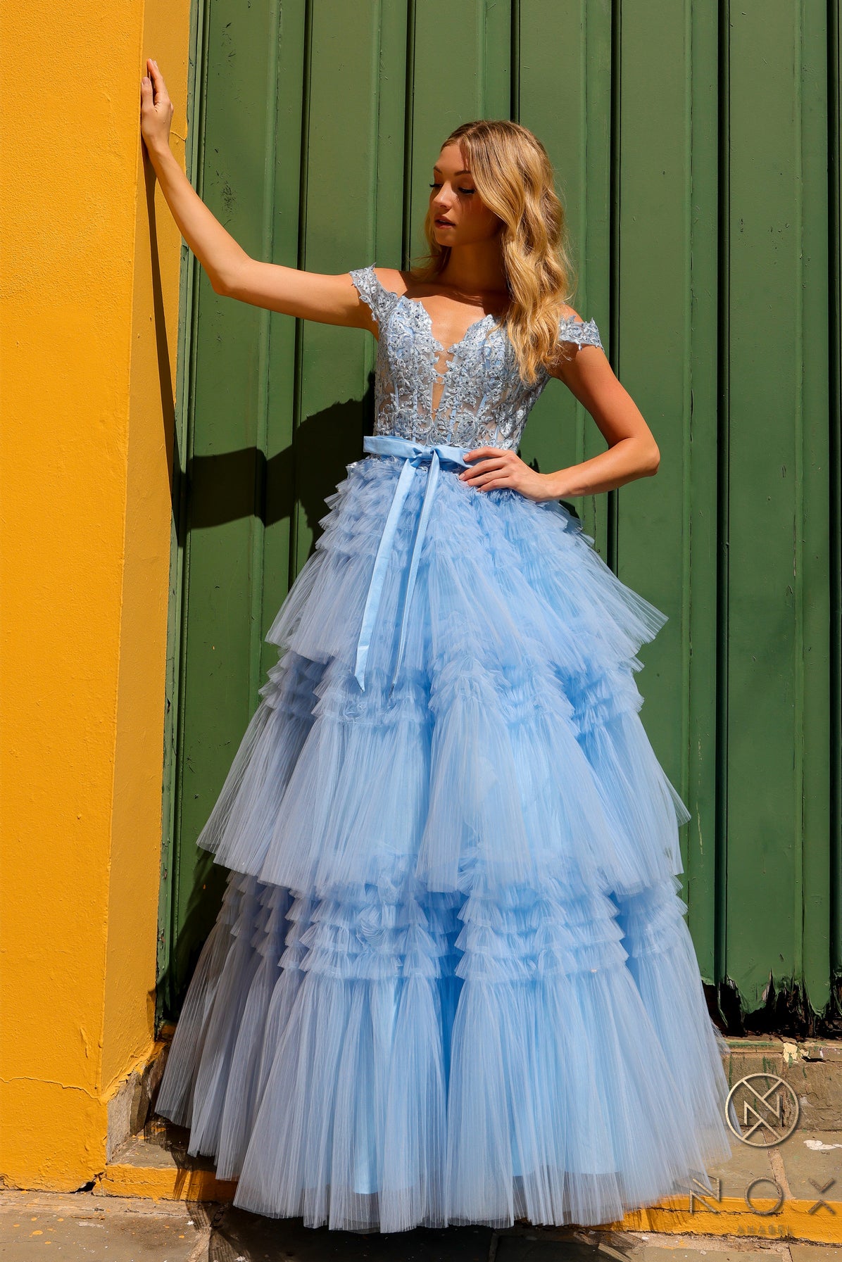 Experience the elegance and sophistication of the Nox Anabel E1293 Prom Dress. With a stunning A-line silhouette, pleated tulle layers, and sheer lace embellished Corset , this dress will make heads turn. Complete with an off-shoulder design and a delicate bow, this dress is perfect for a prom or any formal event.  Sizes: 0-16  Colors: Blue, Mauve, Mint Green, Periwinkle