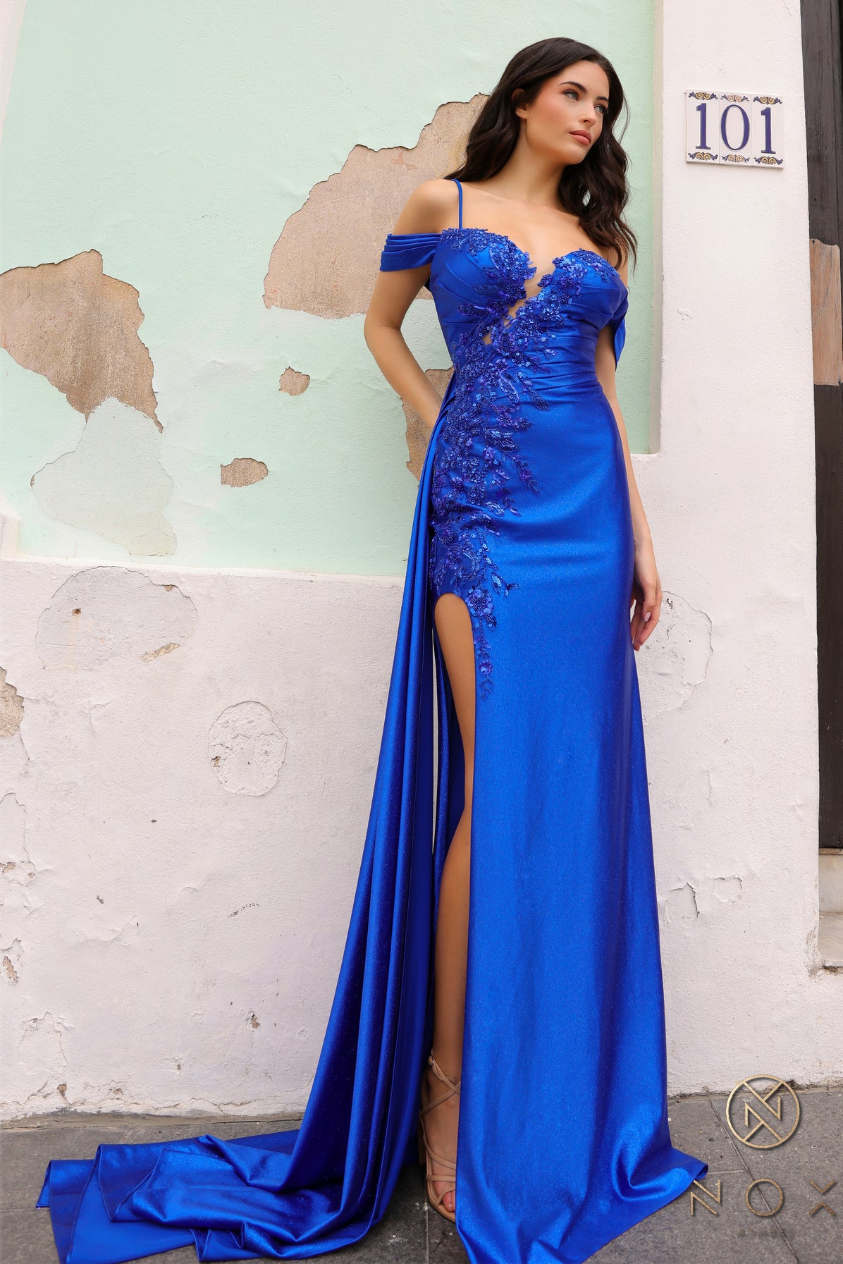 Introducing the Nox Anabel E1451 prom dress, featuring a stunning high slit and off the shoulder lace design. Made with long fitted satin and a sheer illusion bodice, this dress exudes elegance and sophistication. Perfect for prom, pageants, and other formal occasions.  Sizes: 2-16  Colors: Fuchsia, Royal Blue